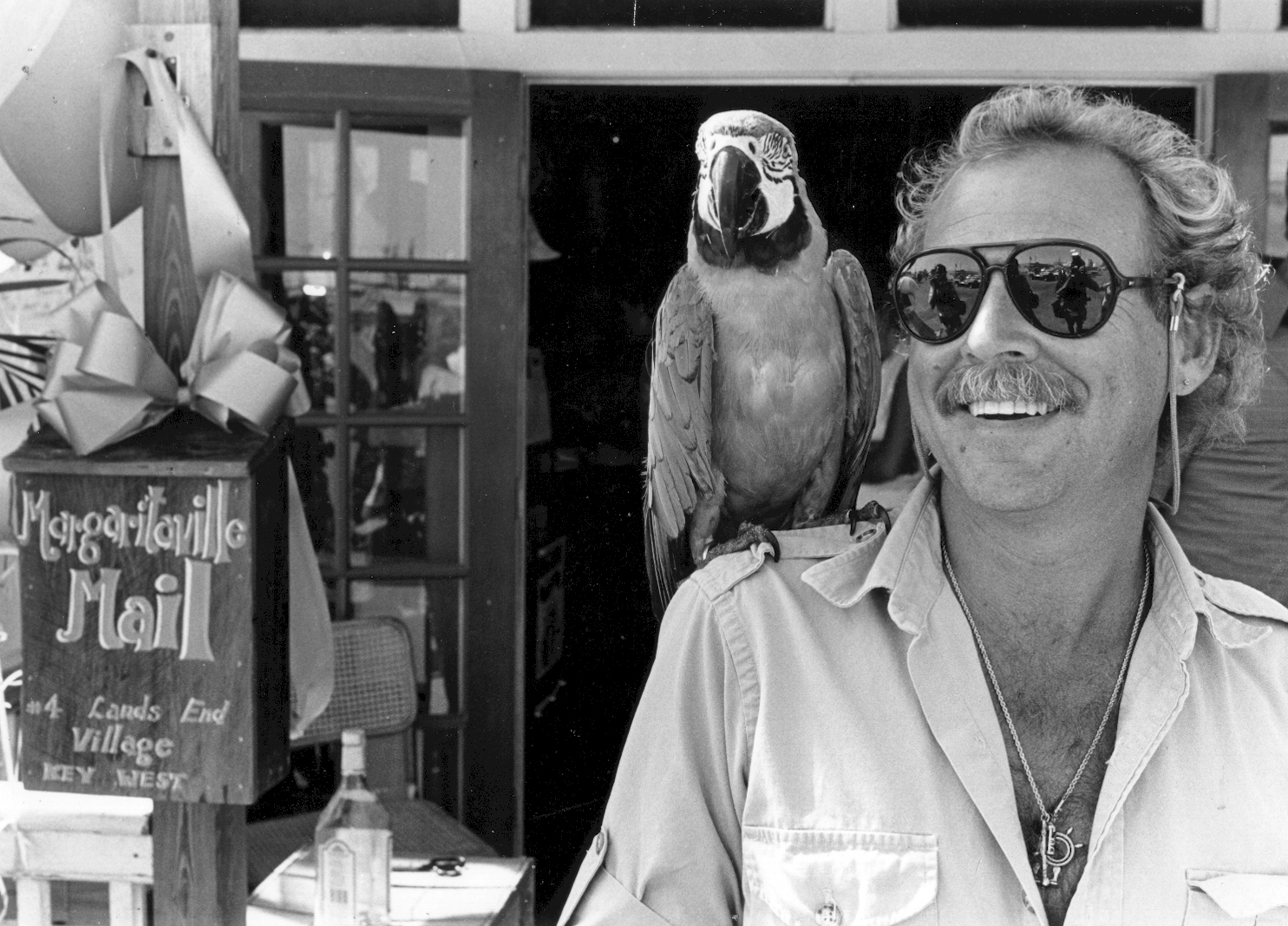 Black and White image of Jimmy Buffet standing outside a Key West novelty shop, smiling with open shirt and wearing large avaitor sunglass with a large parrot perched upon his right shoulder.