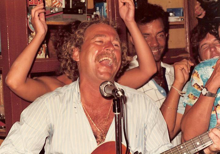 Image of Jimmy Buffett playing guitar and smiling widely while playing to a close-knit group of young people.